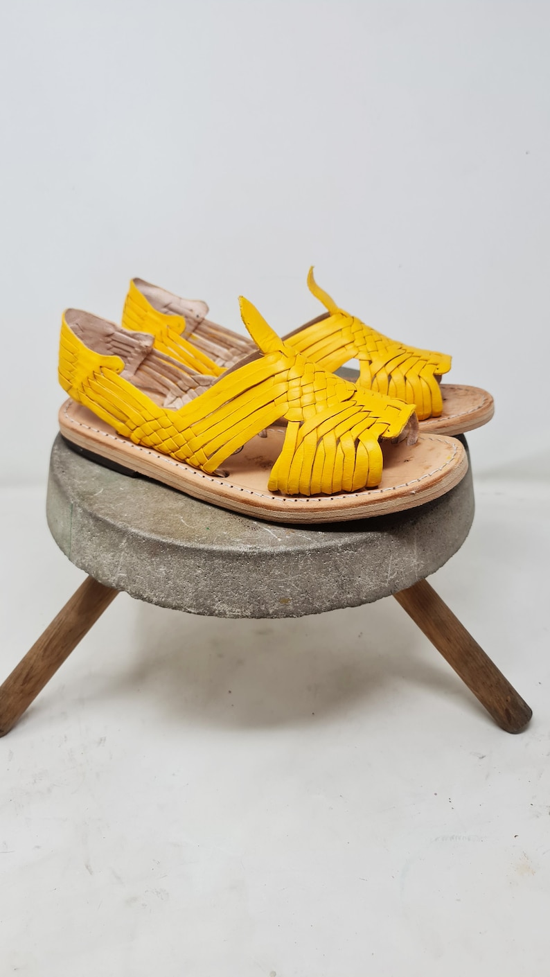 May include: A pair of yellow leather sandals with a woven design. The sandals are sitting on a wooden stool.