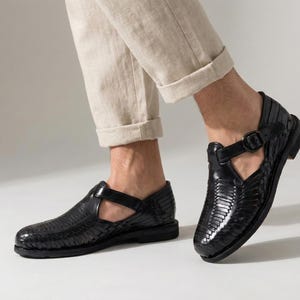 May include: Black leather sandals with a woven design and buckle closure. The sandals have a low heel and are worn with beige pants. The shoes are a stylish and comfortable footwear option.
