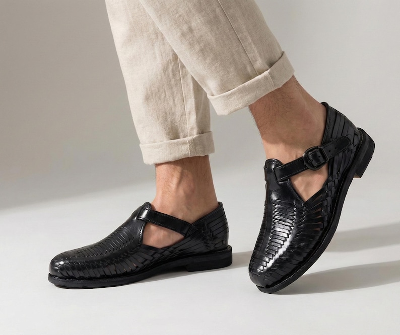 May include: Black leather sandals with a woven design and buckle closure. The sandals have a low heel and are worn with beige pants. The shoes are a stylish and comfortable footwear option.