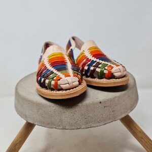 May include: A pair of colorful woven leather sandals with a white base. The sandals are sitting on a concrete pedestal with wooden legs.