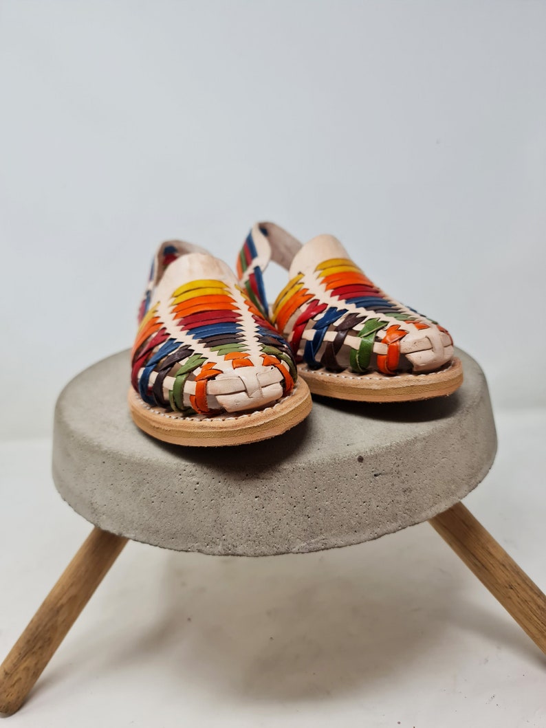 May include: A pair of colorful woven leather sandals with a white base. The sandals are sitting on a concrete pedestal with wooden legs.
