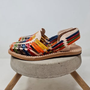 May include: A pair of colorful leather sandals with a woven design. The sandals are on a concrete pedestal with wooden legs.