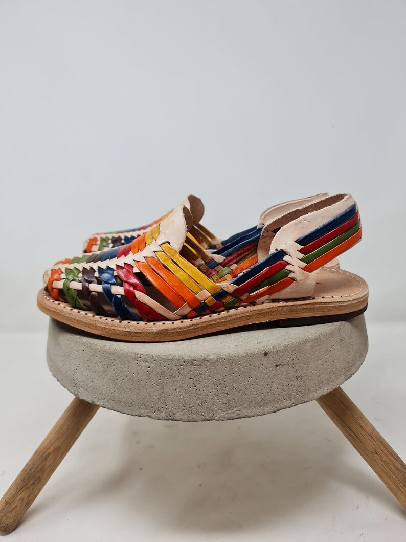 May include: A pair of colorful leather sandals with a woven design. The sandals are on a concrete pedestal with wooden legs.