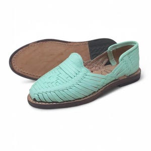 May include: Mint green leather huaraches with woven straps. The flat sole is brown with a stitched edge. The second shoe is upside down, showing the sole. The shoes are a casual style.