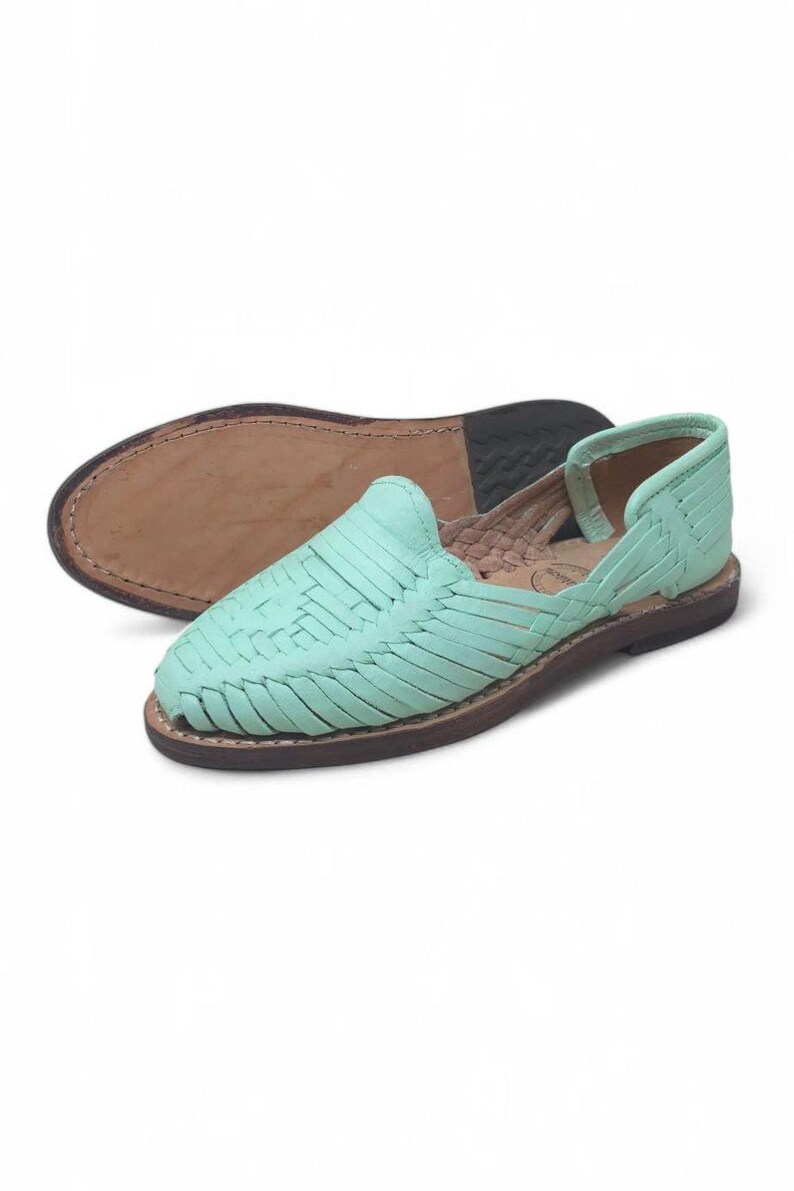 May include: Mint green leather huaraches with woven straps. The flat sole is brown with a stitched edge. The second shoe is upside down, showing the sole. The shoes are a casual style.