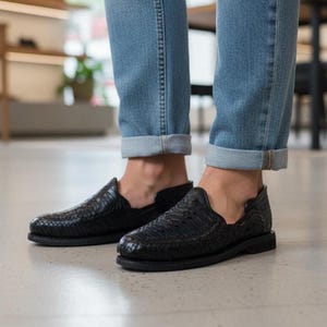 May include: Black woven leather slip-on shoes. The shoes have a textured design and a flat sole. The image shows the shoes paired with blue jeans. The shoes appear to be new and unworn.