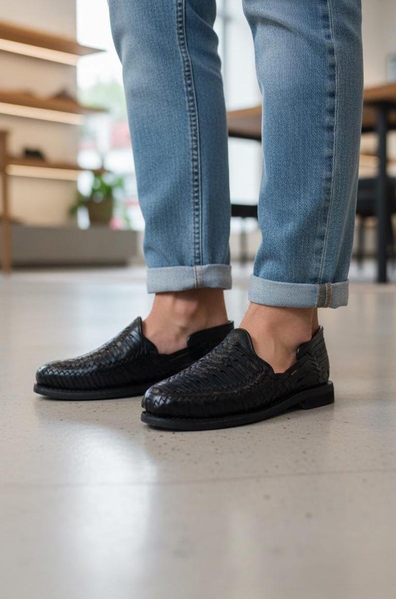 May include: Black woven leather slip-on shoes. The shoes have a textured design and a flat sole. The image shows the shoes paired with blue jeans. The shoes appear to be new and unworn.