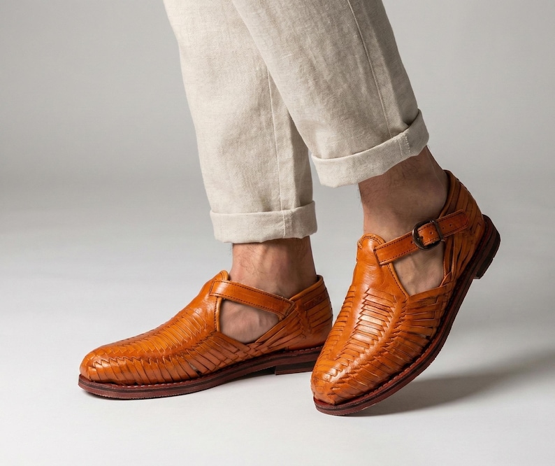 May include: A pair of woven leather sandals in a warm, reddish-brown hue. The sandals feature a T-strap design with a buckle closure and a textured pattern. The soles are a darker shade of brown.