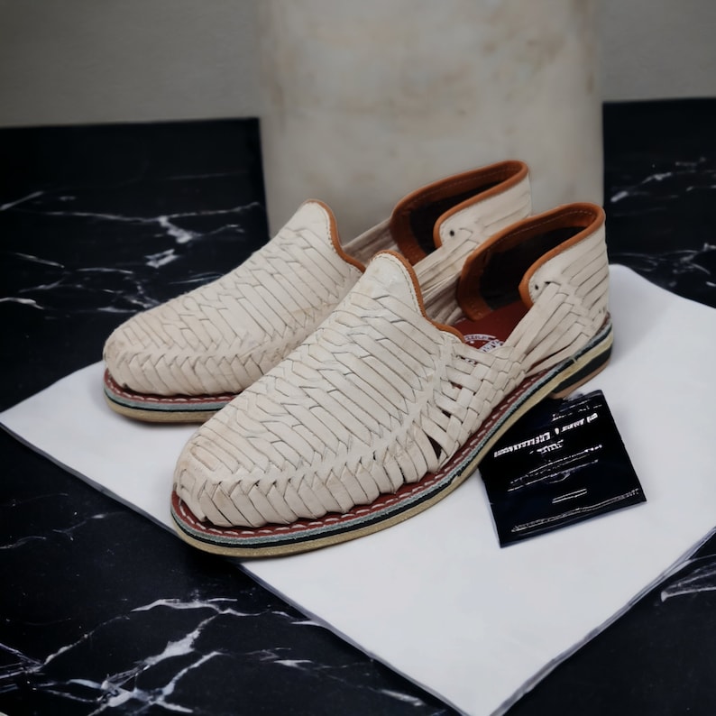 Mocasines de cuero color crema, trenzados a mano, con suela de esparto; destacan por su diseño artesanal sobre fondo de mármol negro, ideal para un estilo sofisticado y natural.