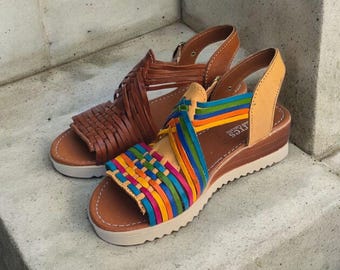 May include: Two pairs of leather sandals. One pair is woven brown leather. The other pair has a colourful woven design with rainbow stripes. Both pairs have a white sole and a small heel.