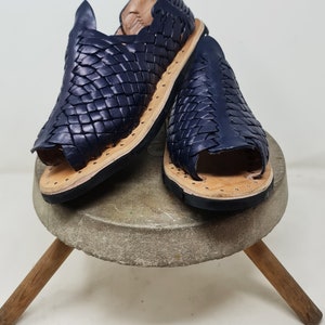 Pu&ograve; includere: Un paio di sandali in pelle blu navy con un design intrecciato. I sandali sono su un supporto in legno.