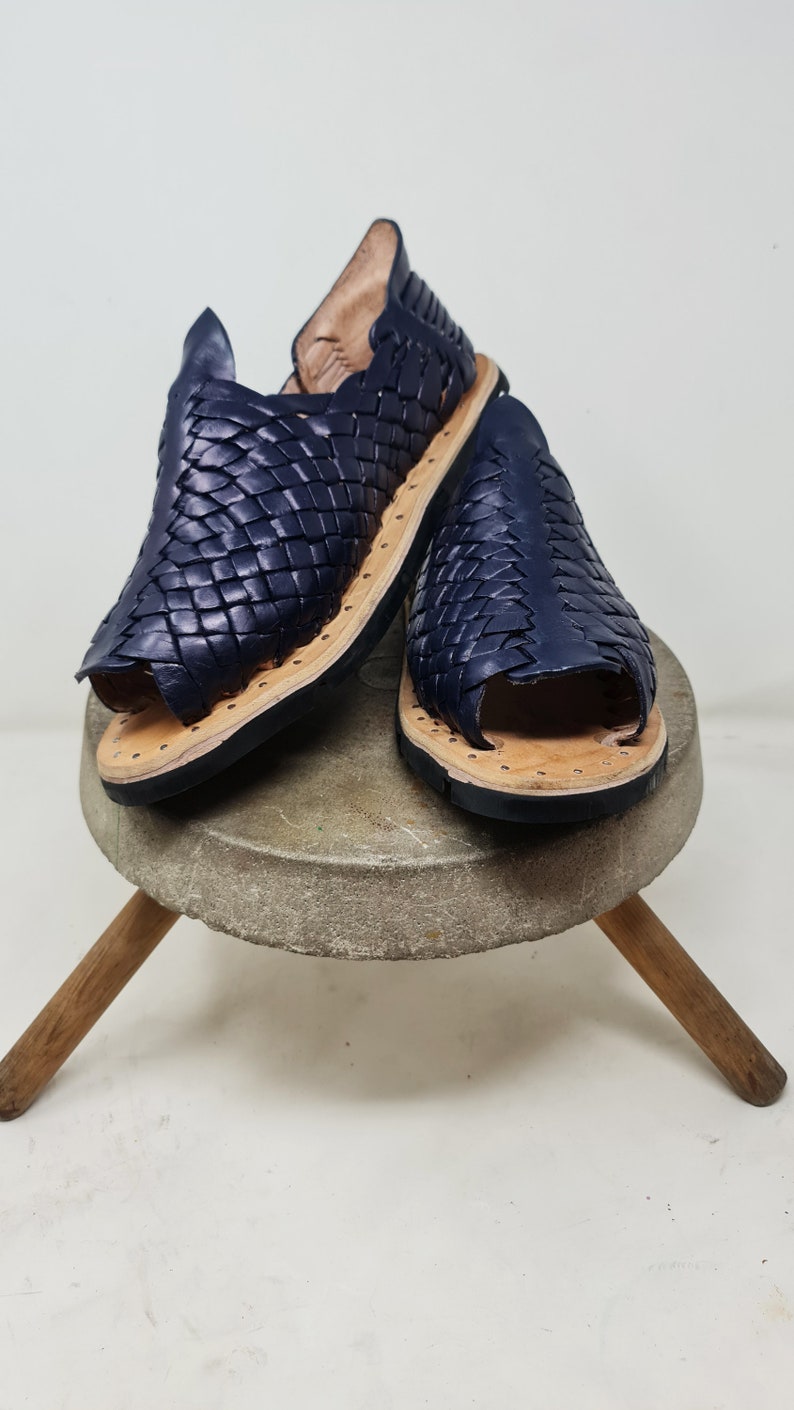 Pu&ograve; includere: Un paio di sandali in pelle blu navy con un design intrecciato. I sandali sono su un supporto in legno.