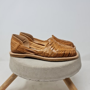 Puede incluir: Un par de sandalias huaraches de cuero color canela con un dise&ntilde;o tejido. Las sandalias se exhiben sobre un peque&ntilde;o taburete redondo de hormig&oacute;n gris con patas de madera. Las sandalias tienen suela plana y dise&ntilde;o abierto.