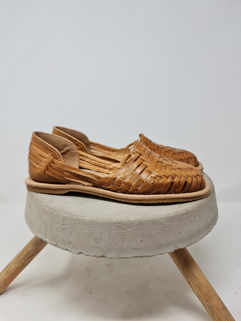 Puede incluir: Un par de sandalias huaraches de cuero color canela con un dise&ntilde;o tejido. Las sandalias se exhiben sobre un peque&ntilde;o taburete redondo de hormig&oacute;n gris con patas de madera. Las sandalias tienen suela plana y dise&ntilde;o abierto.