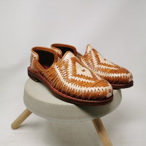 K&ouml;nnte beinhalten: Ein Paar handgefertigte Huaraches aus Leder in einem hellbraunen und cremefarbenen Webmuster. Die Schuhe werden auf einem kleinen, runden, grauen Betonhocker mit Holzbeinen vor wei&szlig;em Hintergrund pr&auml;sentiert. Die Sohlen sind dunkelbraun.
