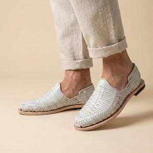 May include: Light gray woven leather shoes with a low profile and a natural-colored sole. The shoes have a slip-on design and are shown from a low angle, highlighting the intricate weave pattern. The shoes are paired with light-colored pants.