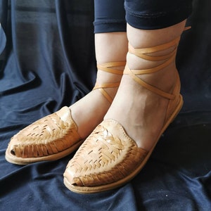 Pu&ograve; includere: Sandali in pelle color cuoio con cinturini intrecciati e lacci che si legano alle caviglie. I sandali sono su uno sfondo di tessuto scuro.