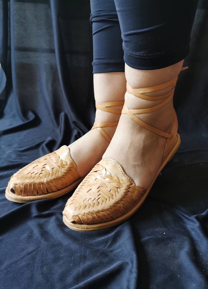 Pu&ograve; includere: Sandali in pelle color cuoio con cinturini intrecciati e lacci che si legano alle caviglie. I sandali sono su uno sfondo di tessuto scuro.