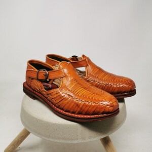 May include: A pair of handcrafted, open-toe leather sandals in a warm, tan color. The sandals feature a woven design across the top and a buckle closure. They are displayed on a small, round, concrete stool with wooden legs.
