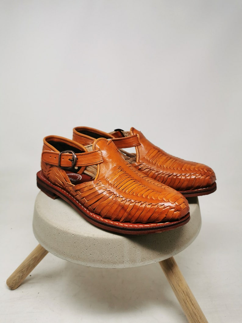 May include: A pair of handcrafted, open-toe leather sandals in a warm, tan color. The sandals feature a woven design across the top and a buckle closure. They are displayed on a small, round, concrete stool with wooden legs.