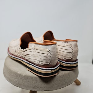 Vista trasera de mocasines de cuero crema trenzados, destacando la textura y la suela multicolor sobre base de concreto, reflejando un diseño artesanal contemporáneo.