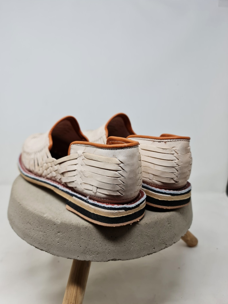 Vista trasera de mocasines de cuero crema trenzados, destacando la textura y la suela multicolor sobre base de concreto, reflejando un diseño artesanal contemporáneo.