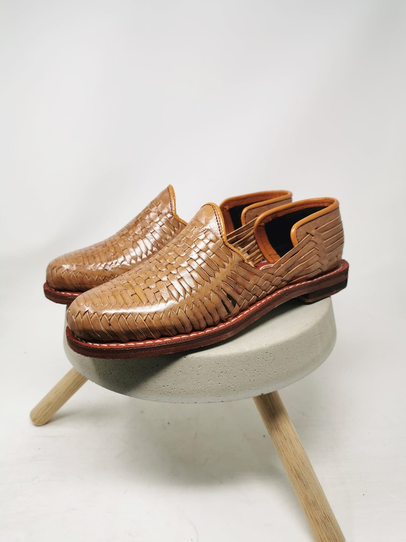 May include: A pair of tan leather woven slip-on shoes with a brown leather sole. The shoes are sitting on a small concrete stool with wooden legs.