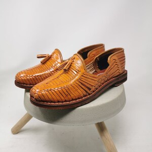 Puede incluir: Un par de sandalias de cuero color canela con un dise&ntilde;o tejido y detalles de borlas. Las sandalias est&aacute;n sobre un peque&ntilde;o taburete de madera.