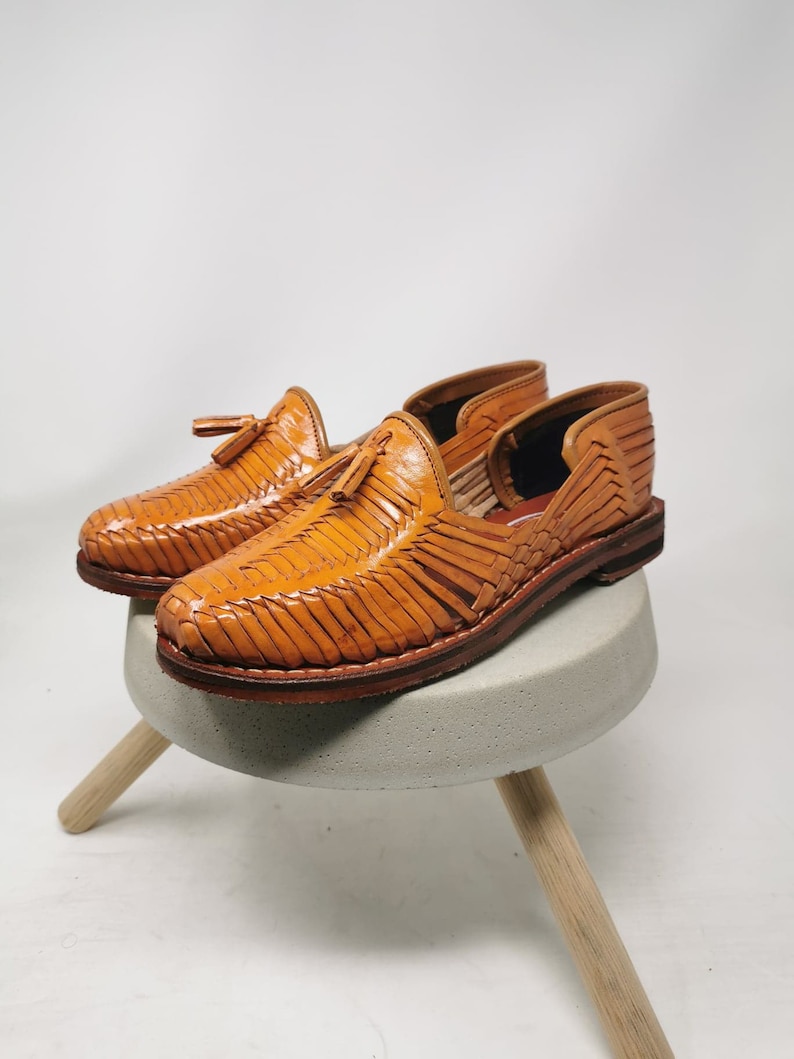 Puede incluir: Un par de sandalias de cuero color canela con un dise&ntilde;o tejido y detalles de borlas. Las sandalias est&aacute;n sobre un peque&ntilde;o taburete de madera.
