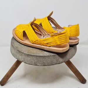 May include: A pair of yellow leather sandals with a woven design. The sandals are sitting on a wooden stool.