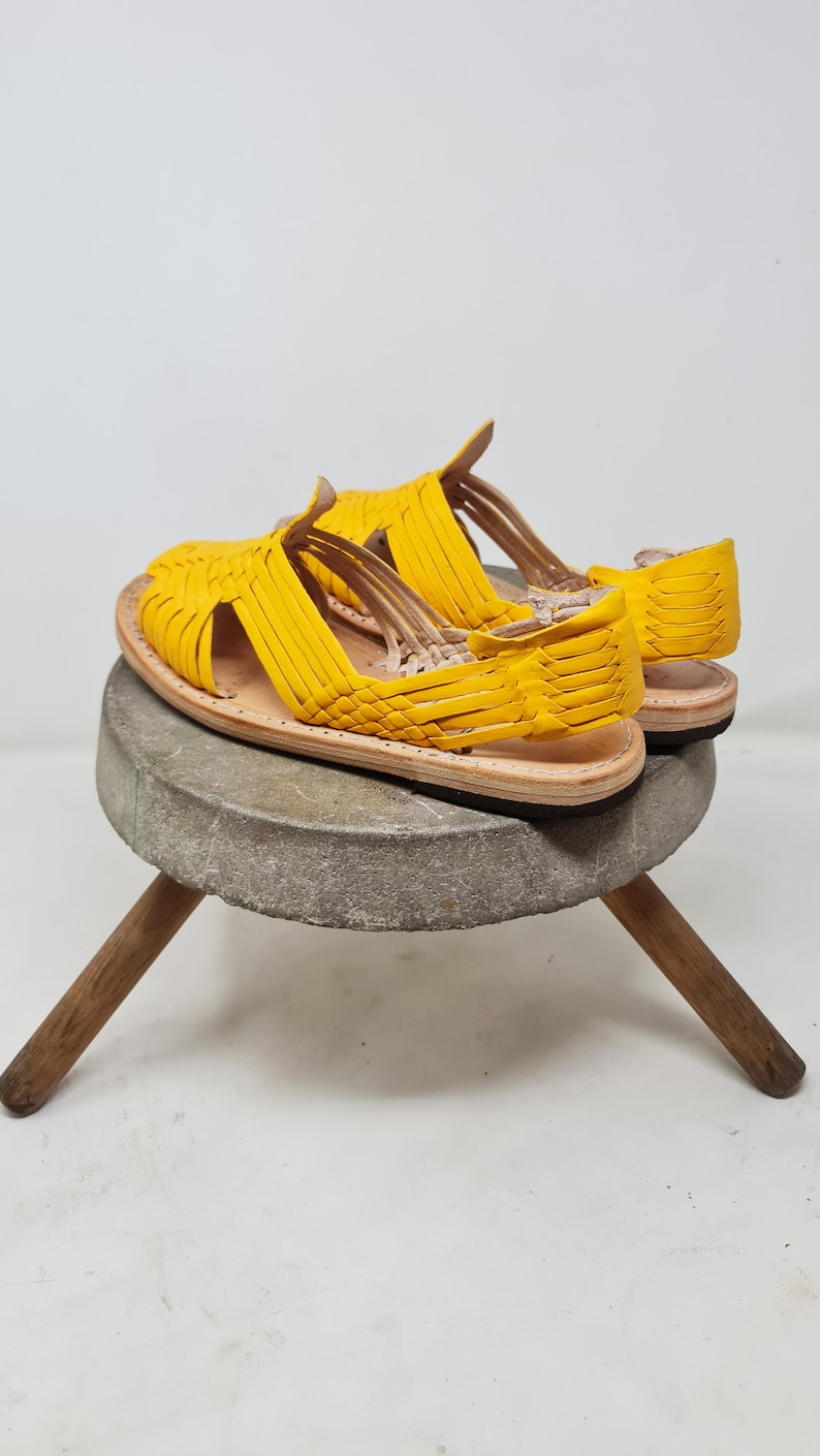 May include: A pair of yellow leather sandals with a woven design. The sandals are sitting on a wooden stool.