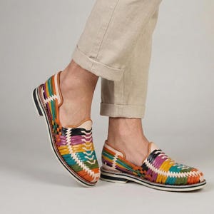 May include: Colorful woven leather shoes with a flat sole. The shoes feature a vibrant pattern of orange, green, purple, and yellow. The shoes are worn with beige pants.