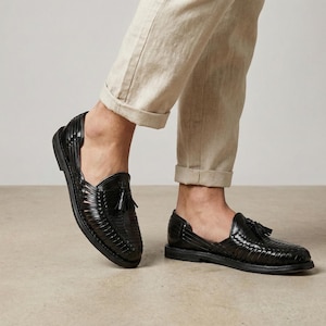 May include: Black leather woven slip-on shoes with a tassel detail. The shoes have a flat sole and a classic design. The shoes are worn with beige pants.