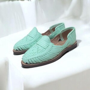 May include: A pair of mint green woven leather sandals with a brown sole. The sandals have a slip-on design with a closed toe and open sides. The background is a white surface with green foliage.