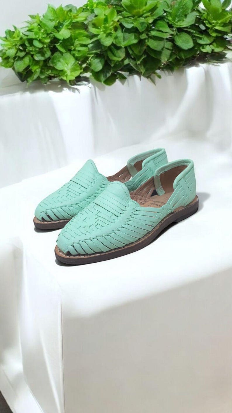 May include: A pair of mint green woven leather sandals with a brown sole. The sandals have a slip-on design with a closed toe and open sides. The background is a white surface with green foliage.