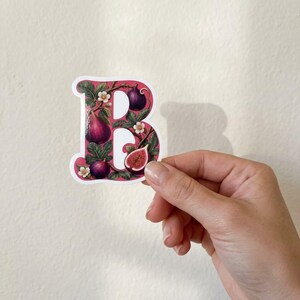 May include: A die-cut sticker in the shape of the capital letter B, featuring a botanical illustration of dark purple figs, green leaves, and small white flowers against a white background, held up against a plain wall.