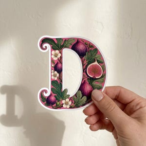 Könnte beinhalten: Ein gestanzter Aufkleber in Form des Großbuchstabens D mit einer botanischen Illustration aus violetten Feigen, grünen Blättern und kleinen weißen Blüten auf rosa Hintergrund, umrandet von einem weißen Rand entlang der Buchstabenform.