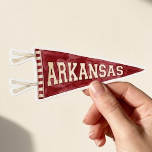 May include: A triangular pennant sticker featuring a deep red watercolour texture with the word ARKANSAS printed in bold, off-white capital letters. The left side displays a vertical striped border in red and white with two attached fabric ties.