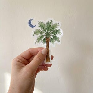 以下が含まれることがあります： 茶色の幹を持つ緑色のヤシの木の透明水彩画が描かれたダイカットステッカー。ヤシの木の左上には、小さな濃い青色の三日月が描かれています。ステッカーには白い縁取りがあり、光沢のある仕上がりになっています。