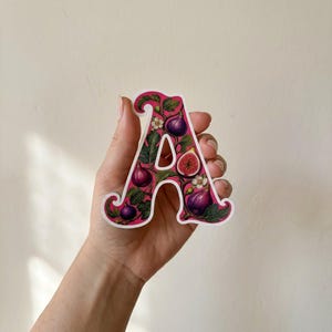 Puede incluir: Una pegatina troquelada con la forma de la letra mayúscula A, que presenta una ilustración botánica de higos morados, hojas verdes y pequeñas flores blancas sobre un fondo rosa vibrante, con un borde blanco que perfila la letra.