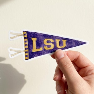 May include: A triangular pennant sticker featuring a purple watercolour texture with the letters LSU in gold. The left side has a vertical gold and purple striped border with two white ties extending outward from the base of the pennant.
