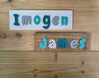 May include: Two rectangular wooden signs featuring raised, colorful letters. The top sign is white with the name Imogen in teal and grey polka-dot letters. The bottom sign is natural wood with the name James in light blue and grey polka-dot letters.