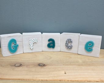 May include: Five white wooden blocks arranged in a row, each featuring a single letter to spell the word grace. The letters G, a, and e are teal with white dots, while the letters r and c are white with teal dots and grey with white dots.