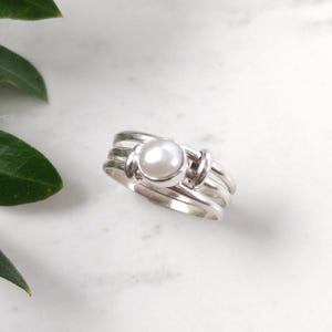 May include: A silver ring with a large, round, white pearl set in the center. The ring has a multi-band design with a knot-like detail on the sides. The ring is on a white marble surface with green leaves in the background.