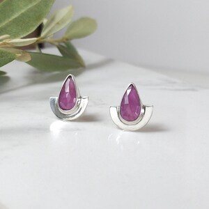 May include: A pair of silver stud earrings. Each earring features a teardrop-shaped, deep pink gemstone set within a silver, half-moon shaped frame. The earrings are displayed on a white surface with a blurred green leaf in the background.