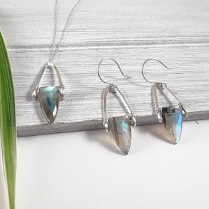 May include: A silver necklace and matching earrings set. The necklace features a teardrop-shaped, iridescent gemstone pendant suspended from a delicate chain. The earrings have a similar design, with the gemstone held in a silver frame.