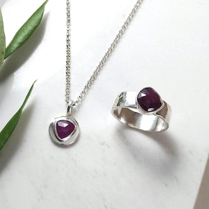 May include: A silver ring and necklace set with a deep purple gemstone. The ring is a wide band with a hammered texture. The necklace has a delicate chain and a pendant with a round bezel setting.