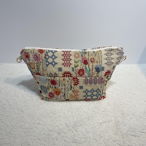 May include: A beige zippered pouch with a floral and geometric pattern. The pouch has two white straps on the sides.