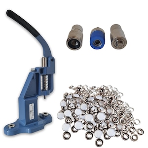 May include: A blue snap press machine with a black handle, three metal attachments, and a pile of silver and white snaps. The machine has a spring mechanism and a small logo on the side. The snaps have a white, circular top.