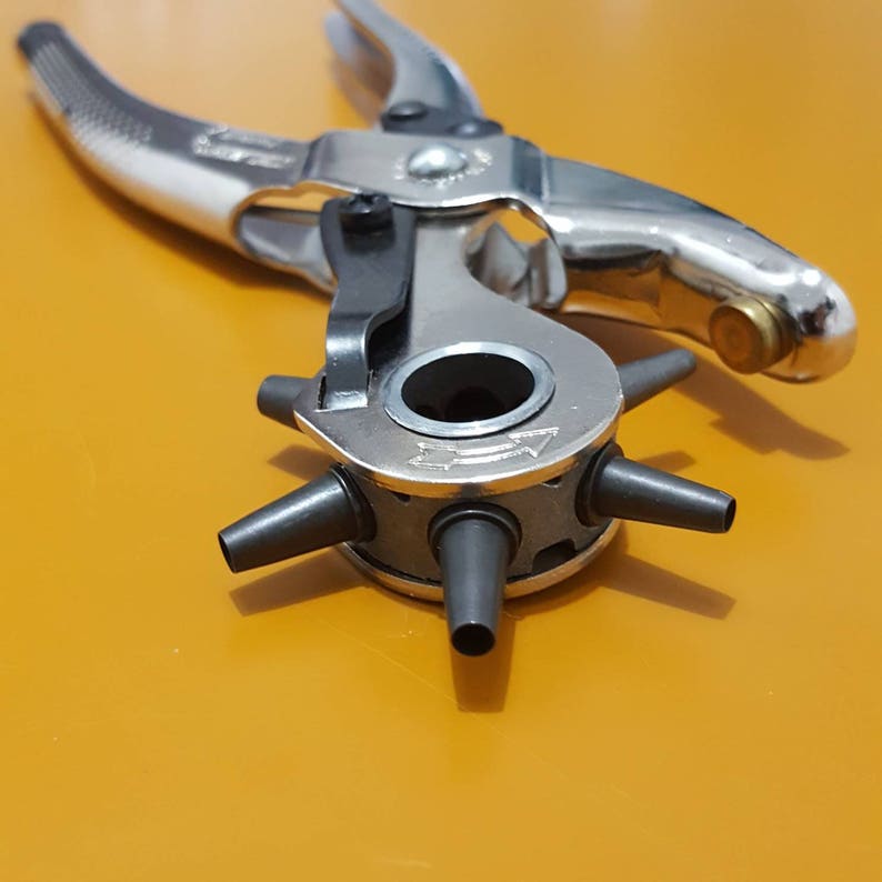 May include: A silver and black multi-size hole punch tool. The tool has a rotating head with multiple sized punches and a spring-loaded handle. The tool is designed for leather, paper, or other materials. The tool is on an orange surface.